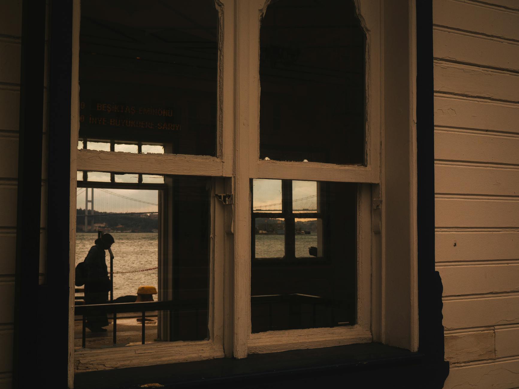 Vista del Bósforo a través de una ventana de muelle, reflejando la silueta de un viajero y un puente distante al atardecer.