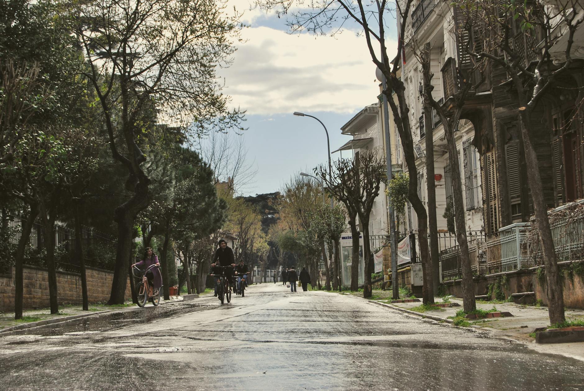 Vista de una calle arbolada, húmeda y con casas antiguas de madera a la derecha, evocando el encanto nostálgico de Büyükada y las Islas de los Príncipes en Estambul. Varias personas disfrutan del paseo en bicicleta, reflejando la vida sin asfalto característica de estas islas turcas.