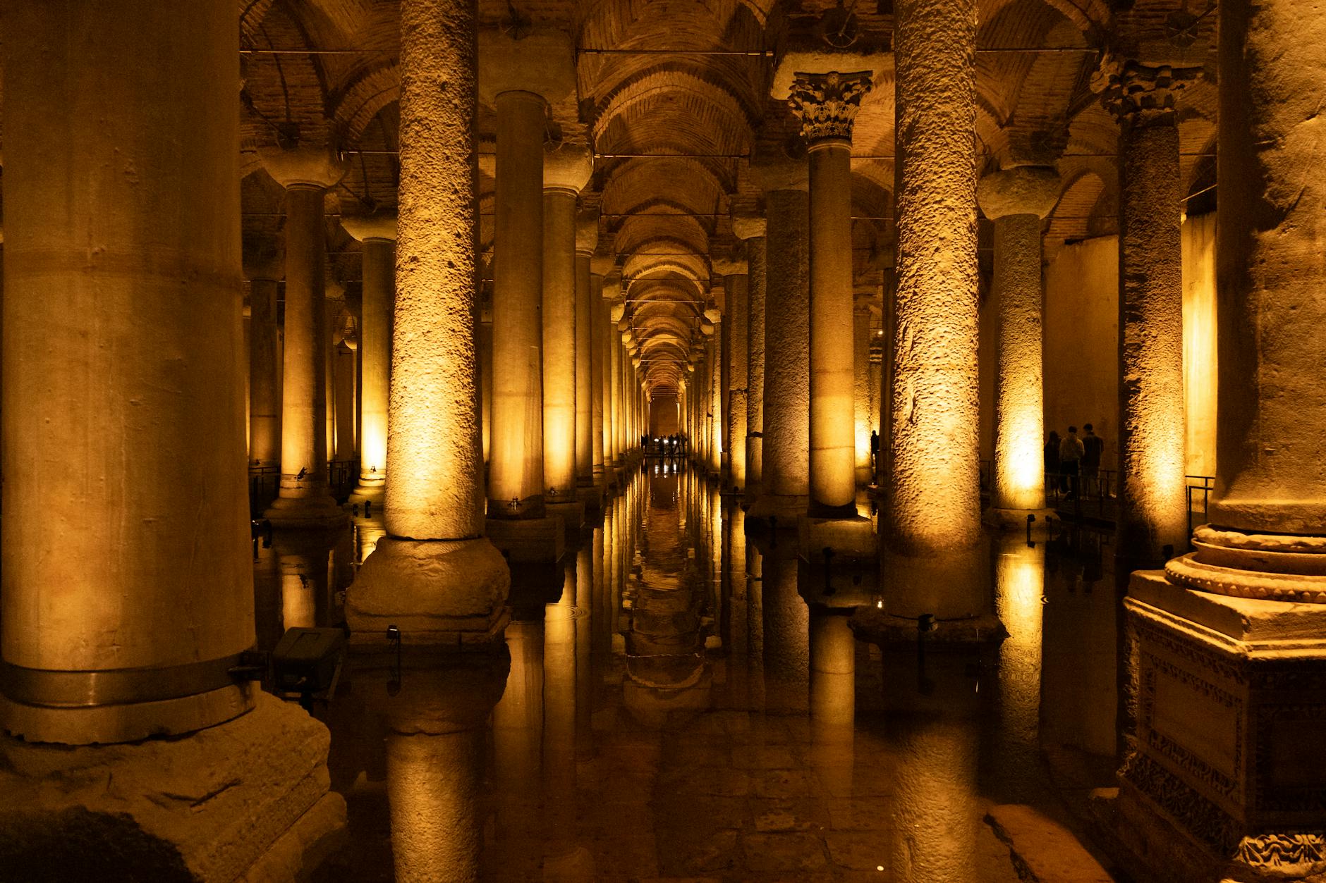 Interior de la Cisterna de Teodosio (Şerefiye) en Estambul, mostrando las largas filas de columnas iluminadas con luz ámbar y sus reflejos sobre el suelo anegado.