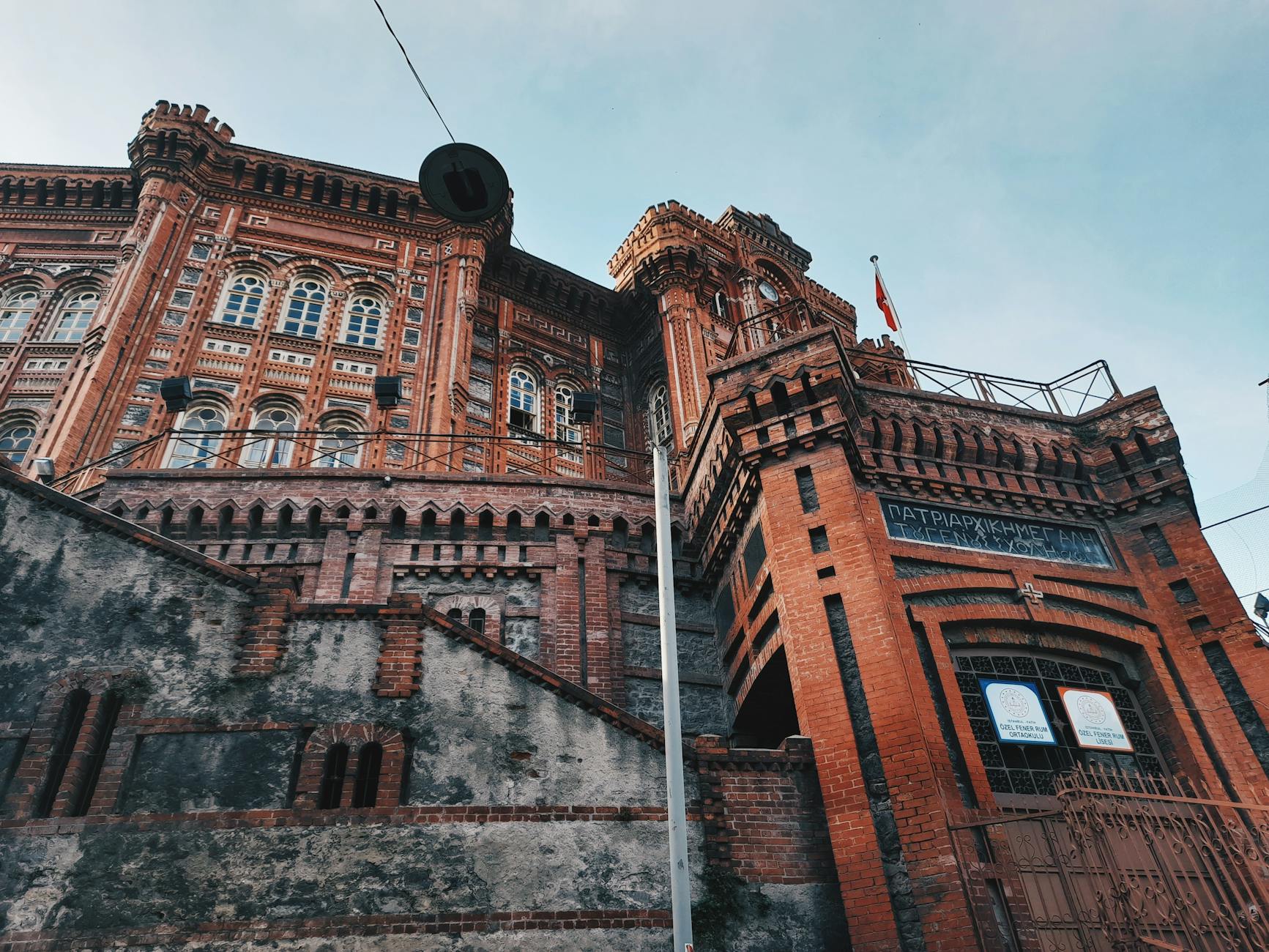 Vista imponente en contrapicado del imponente edificio del Colegio Patriarcal Griego de Fener en Estambul, un gigante rojo de ladrillo visto con detalles neogóticos. La foto captura la empinada cuesta de Fener, haciendo honor al sentimiento de "Me dejo los pulmones en las cuestas de Fener por este gigante rojo" al mostrar su escala majestuosa contra un cielo pálido. Se aprecian inscripciones en griego y los letreros de la escuela en la fachada de ladrillo.