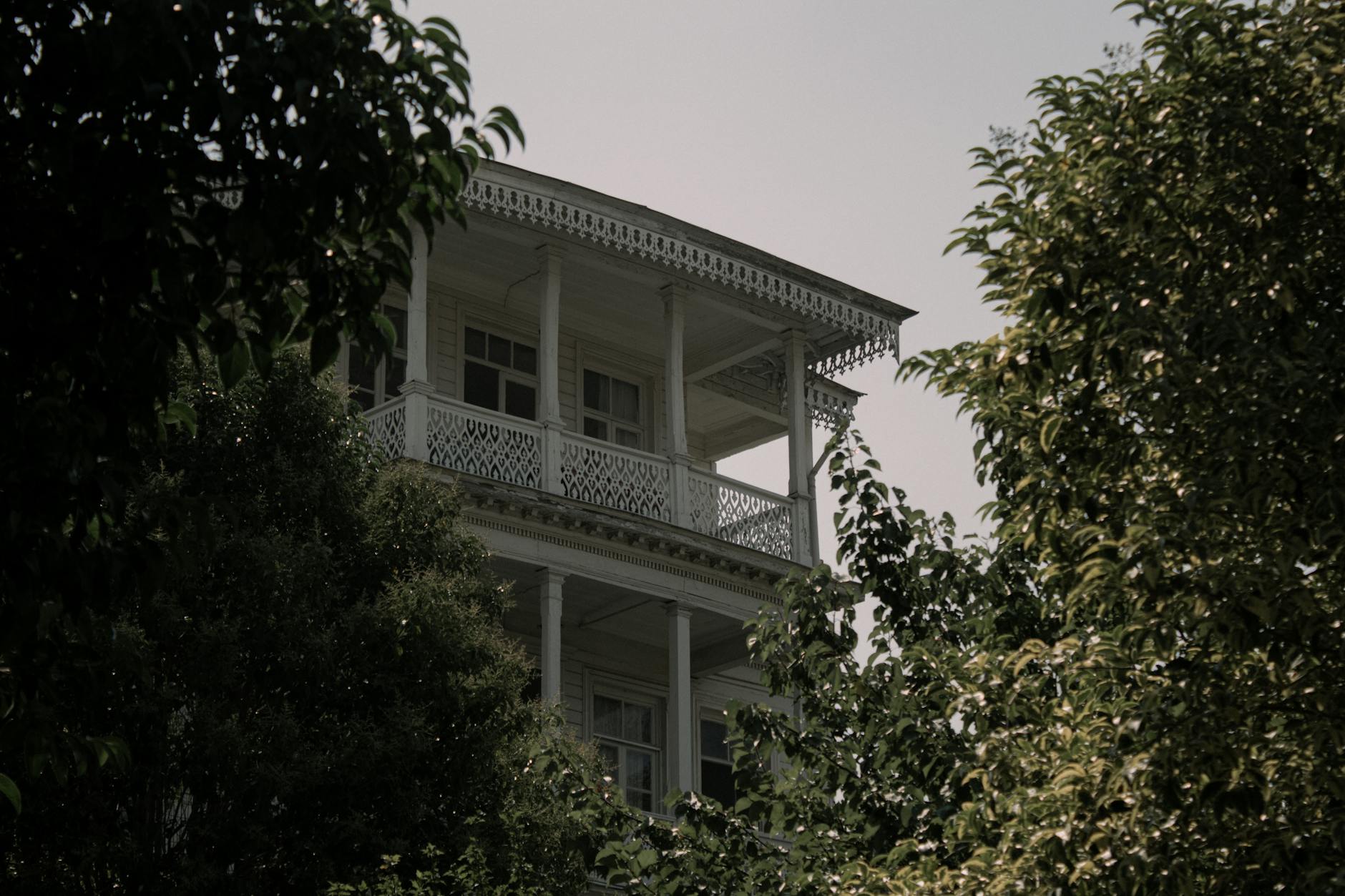 Fachada de una casa otomana de madera blanca con balcones decorativos, oculta parcialmente entre el follaje verde en Kuzguncuk, Estambul.