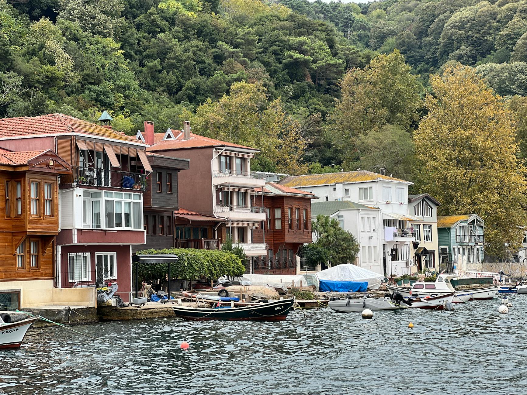 Vista de casas tradicionales de madera y colores a orillas del Bósforo en Kuzguncuk, Estambul, con pequeños botes amarrados y una colina boscosa de fondo.