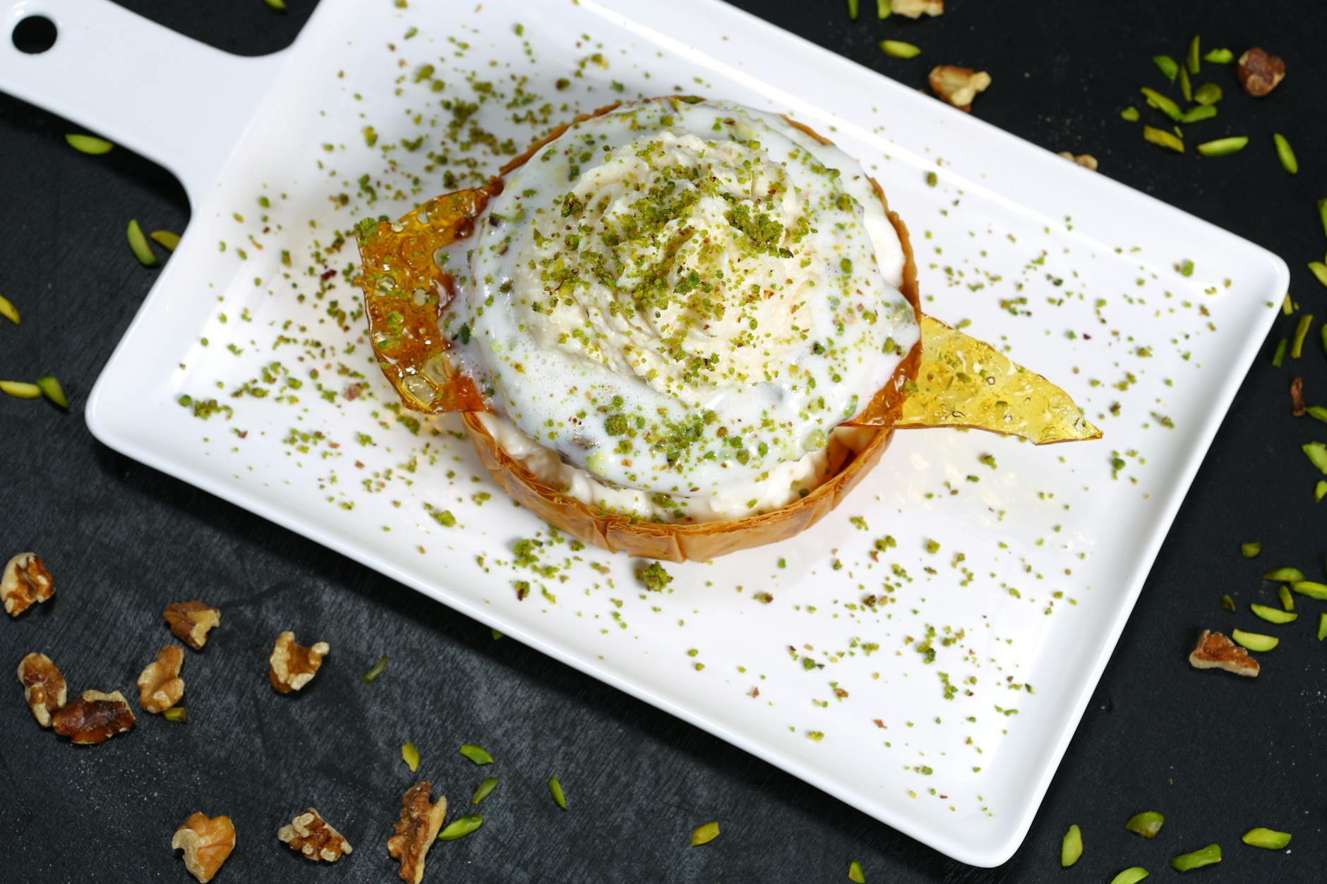 Postre otomano con helado y capas de masa crujiente, cubierto con pistachos molidos y adornado con caramelo, servido en plato blanco.