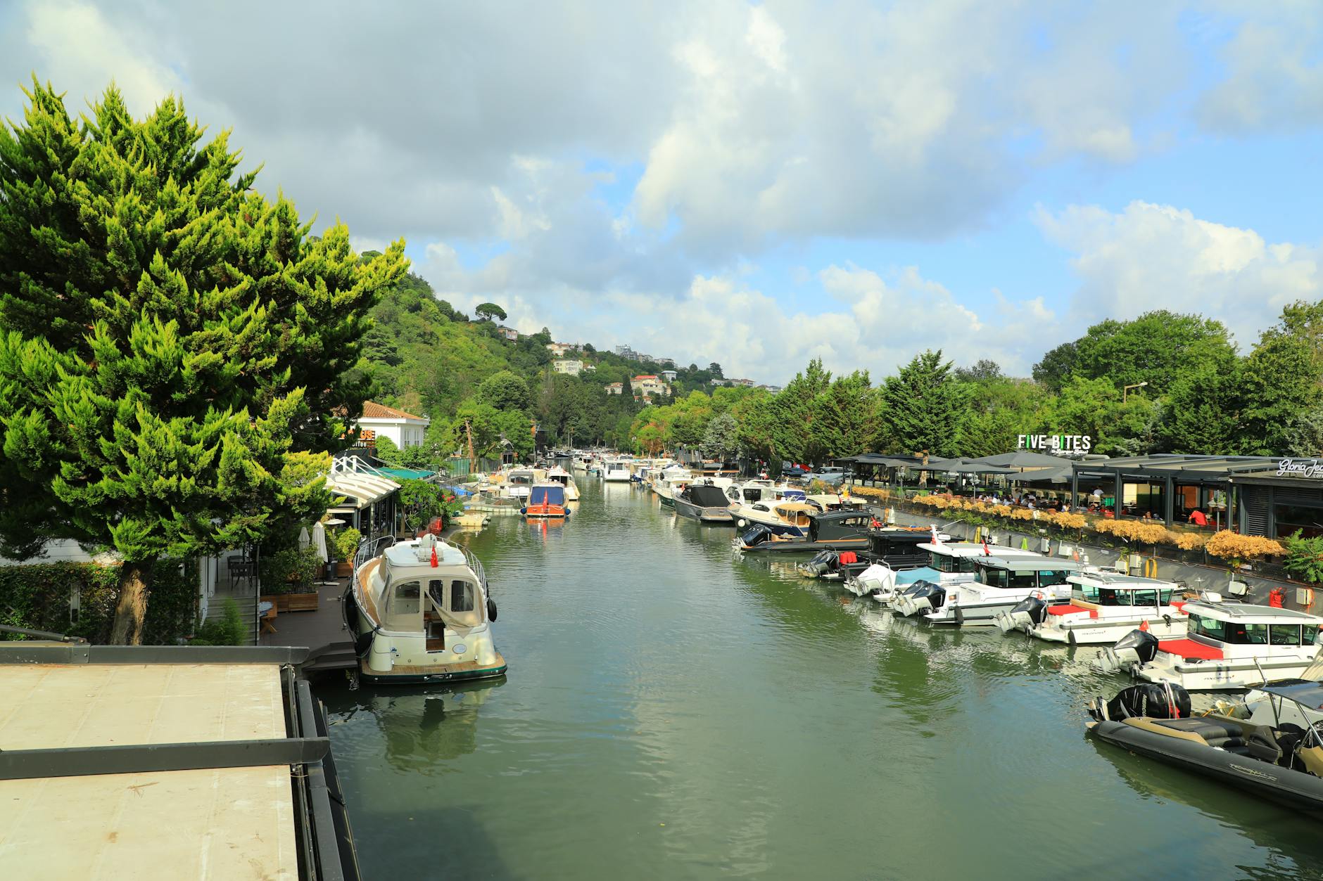 Barcos atracados a lo largo del río Riva con cafeterías y vegetación en las orillas.