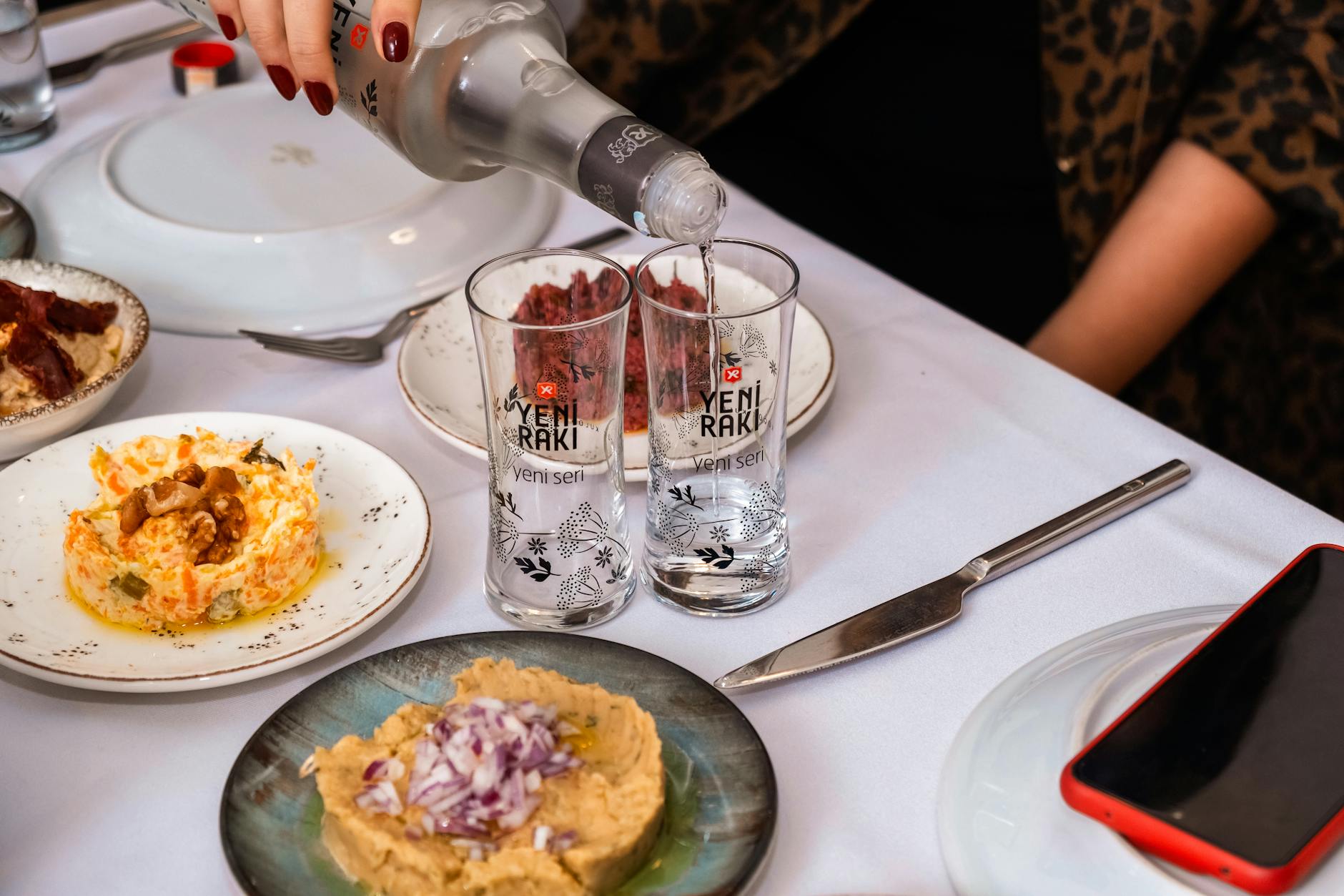 Una persona sirve Yeni Raki en vasos altos acompañados de varios platos de meze turco, ideal para una cena de amigos en Estambul.